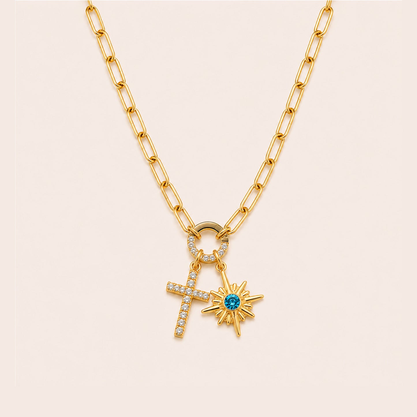A gold chain necklace featuring a double-drop pendant design with a small pave-studded cross and a gold sunburst charm set with a central blue gemstone, displayed on a light cream background.