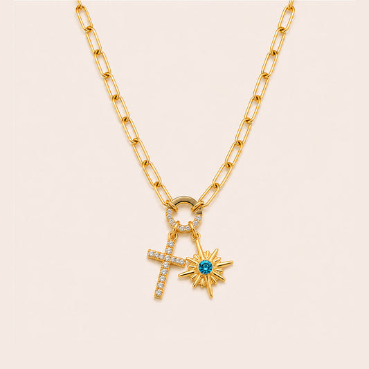 A gold chain necklace featuring a double-drop pendant design with a small pave-studded cross and a gold sunburst charm set with a central blue gemstone, displayed on a light cream background.