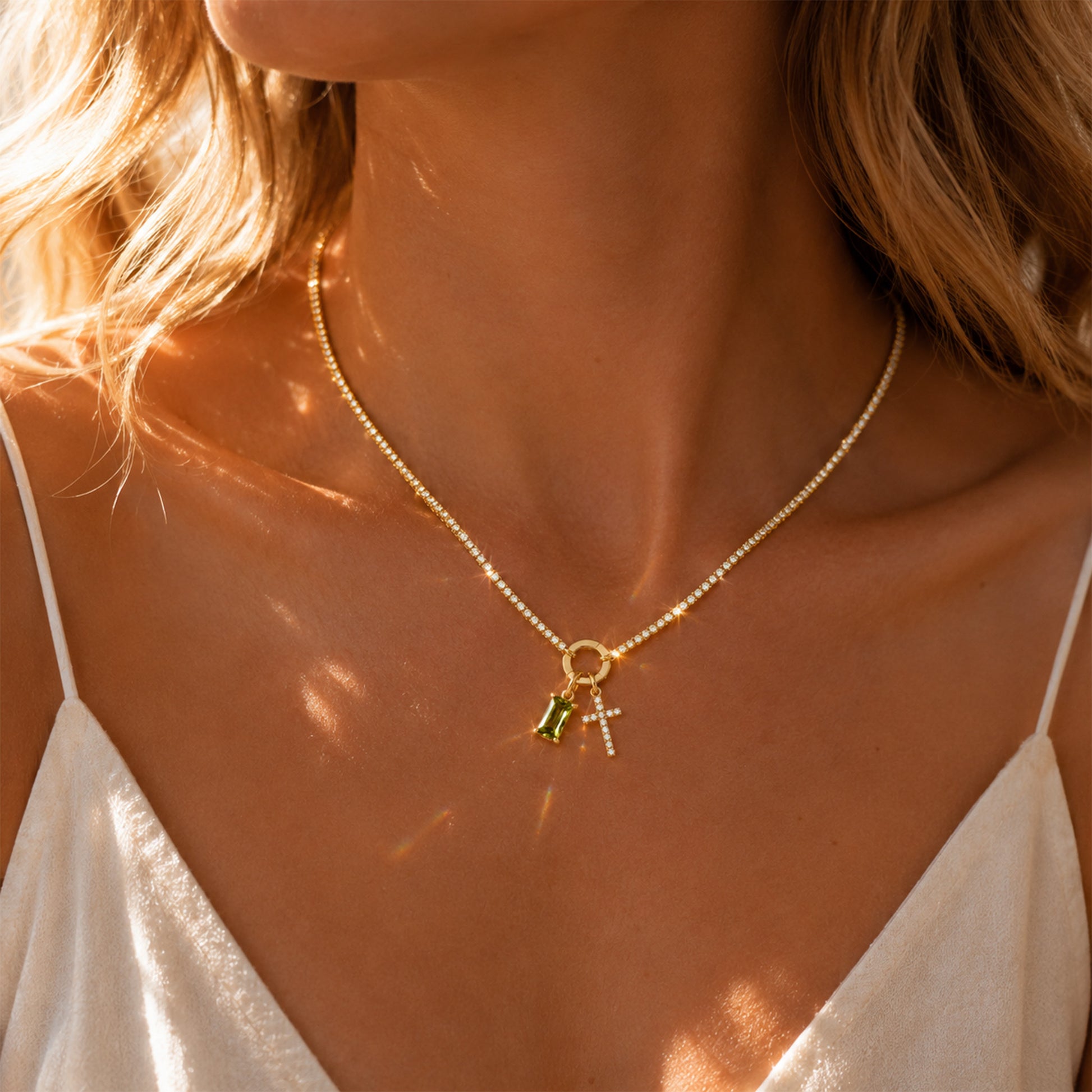 A close-up lifestyle shot of a model wearing a delicate gold chain necklace with a small charm featuring a green heart and a silver cross, styled with a white camisole under warm sunlight.