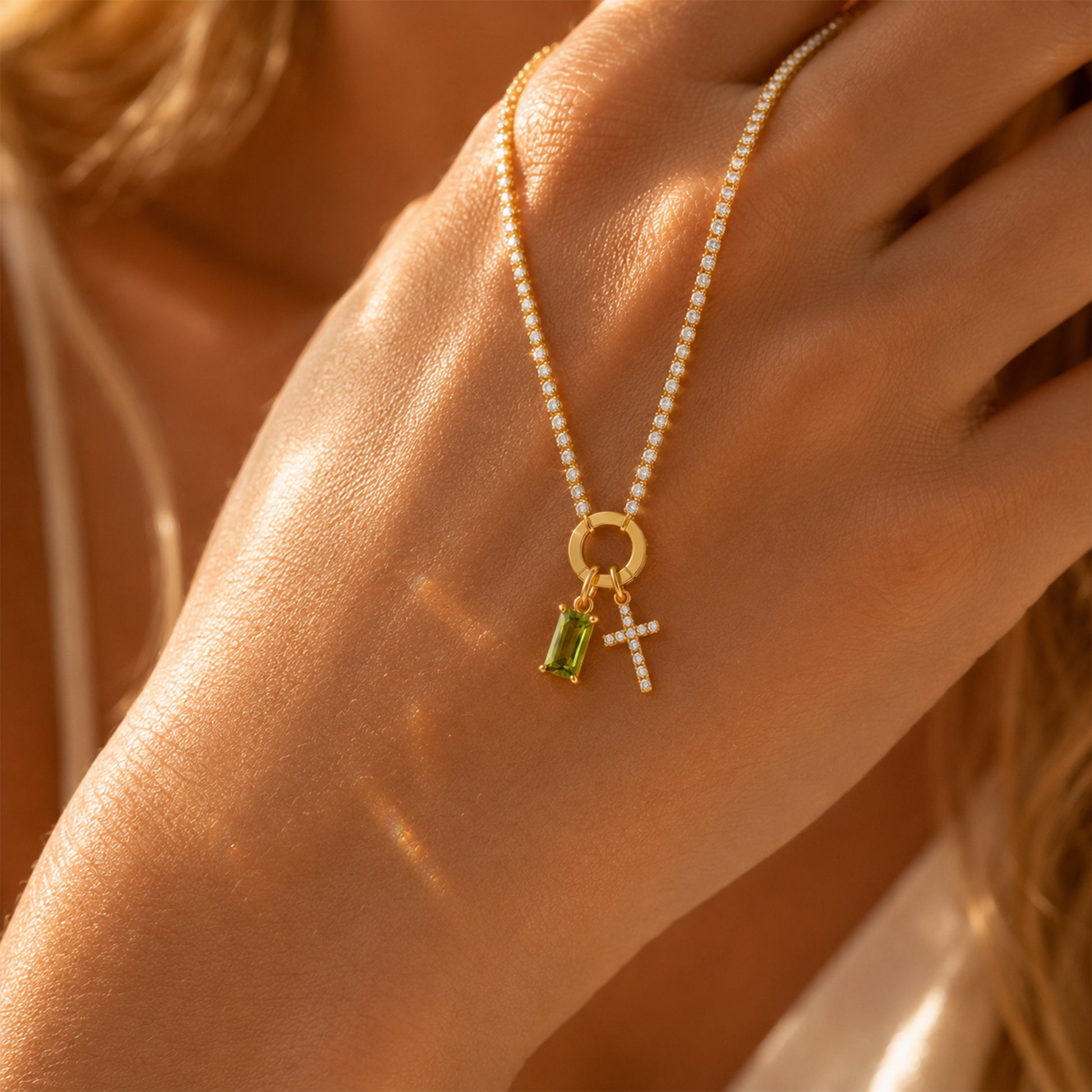 A close-up of a delicate gold necklace with a small ring connector, featuring a green rectangular gemstone charm and a small cross charm, worn on a model's hand.
