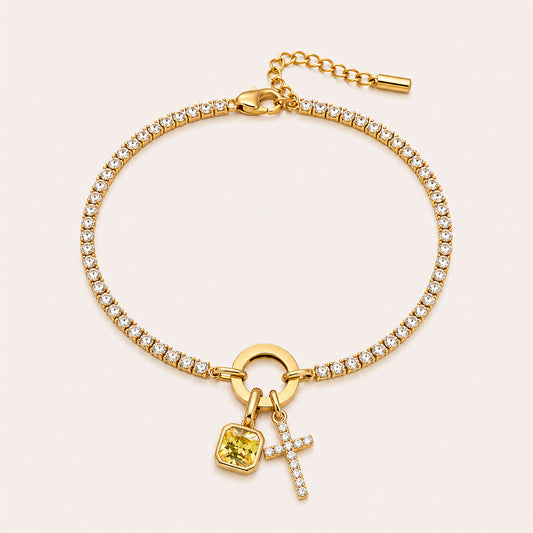A gold-plated bracelet featuring a row of clear pavé-set round crystals, a circular ring connector, and two dangling charms: a square yellow gemstone charm and a small cross charm paved with clear crystals, all on a light pink background.