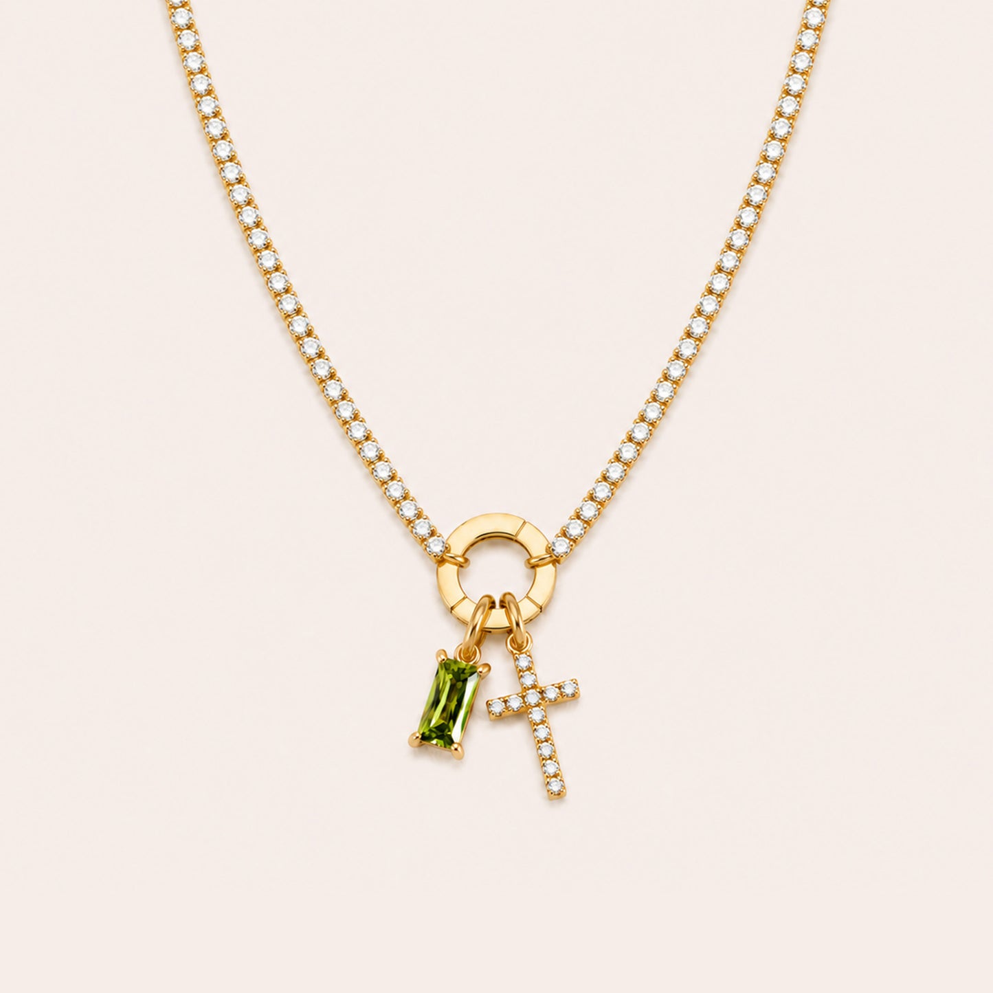 A gold chain necklace featuring a circular ring charm, a small cross charm set with clear stones, and a green rectangular gemstone charm, displayed on a light beige background.