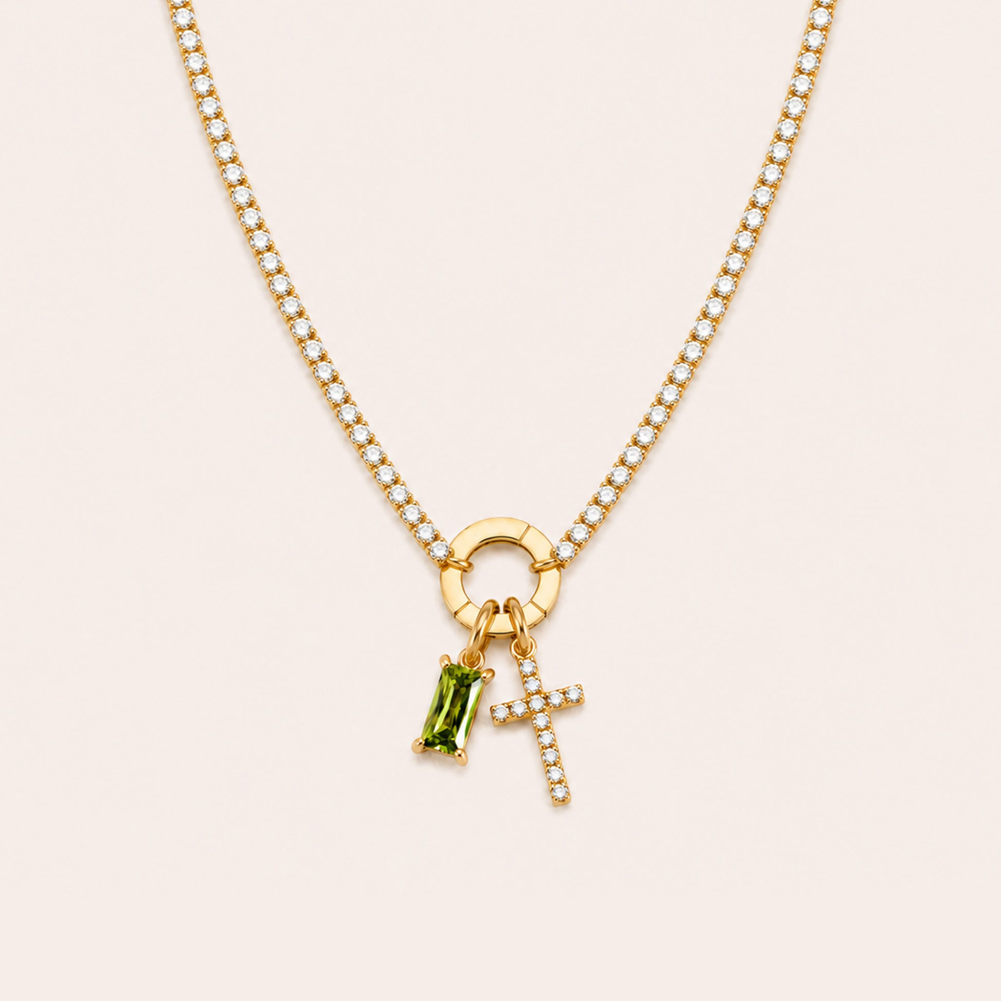 A gold chain necklace featuring a circular ring charm, a small cross charm set with clear stones, and a green rectangular gemstone charm, displayed on a light beige background.