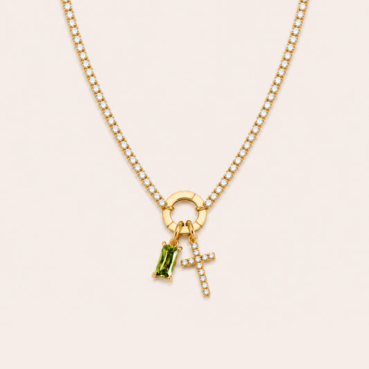 A gold chain necklace featuring a circular ring charm, a small cross charm set with clear stones, and a green rectangular gemstone charm, displayed on a light beige background.