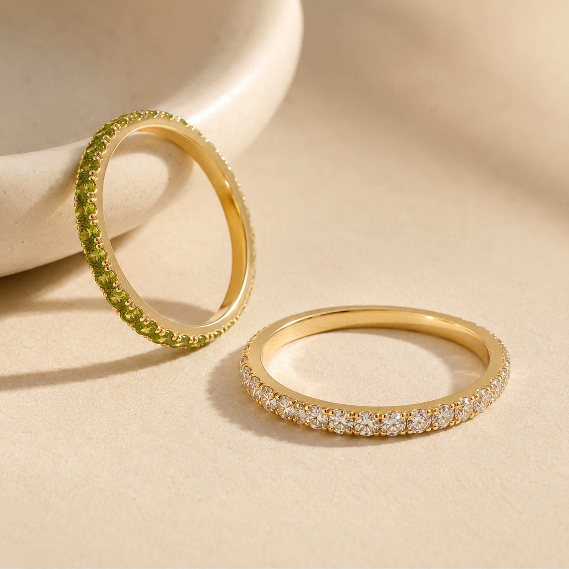 Two gold eternity rings side by side — peridot green birthstone band and white CZ diamond band on stone prop