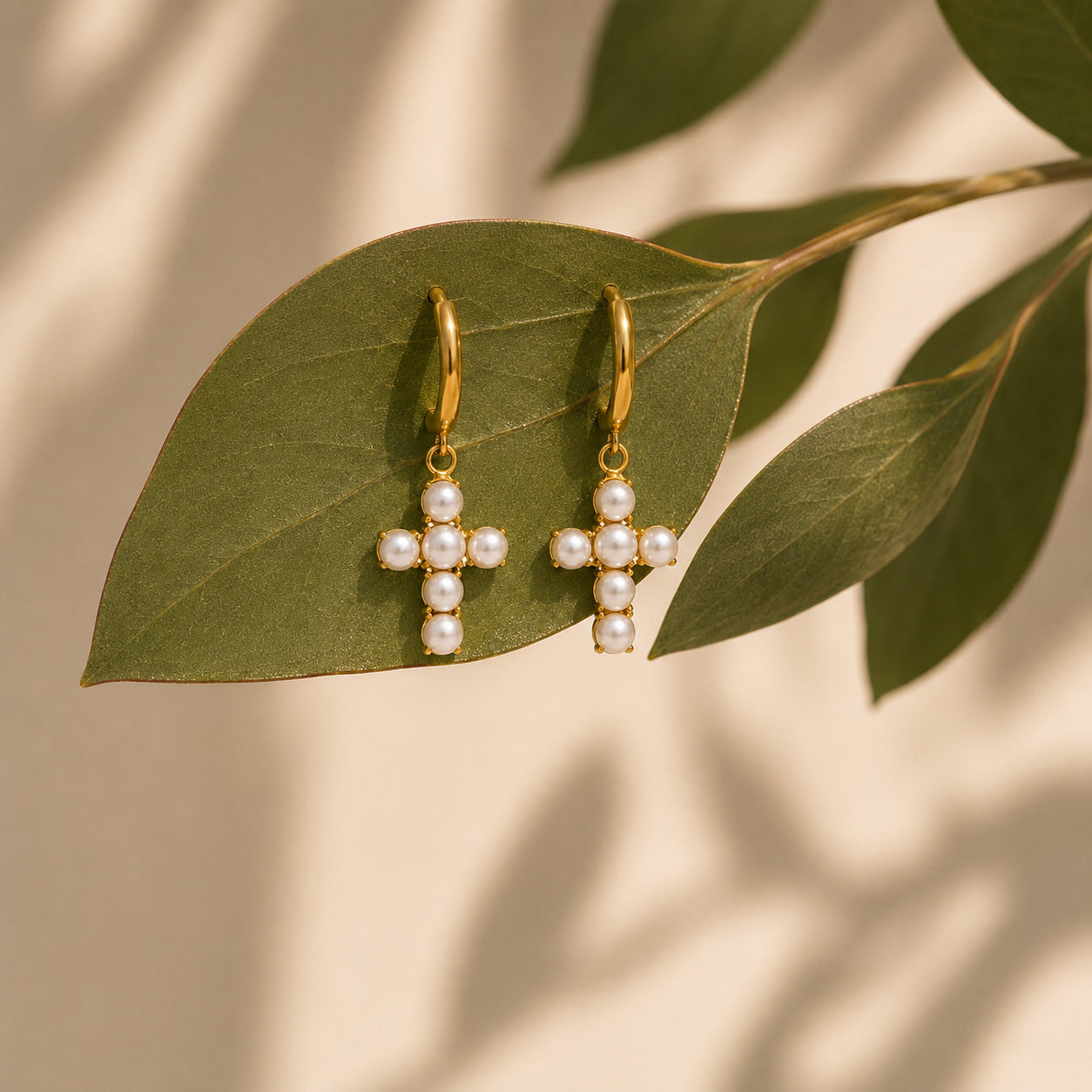 Pair of gold pearl cross drop hoop earrings resting on a green leaf