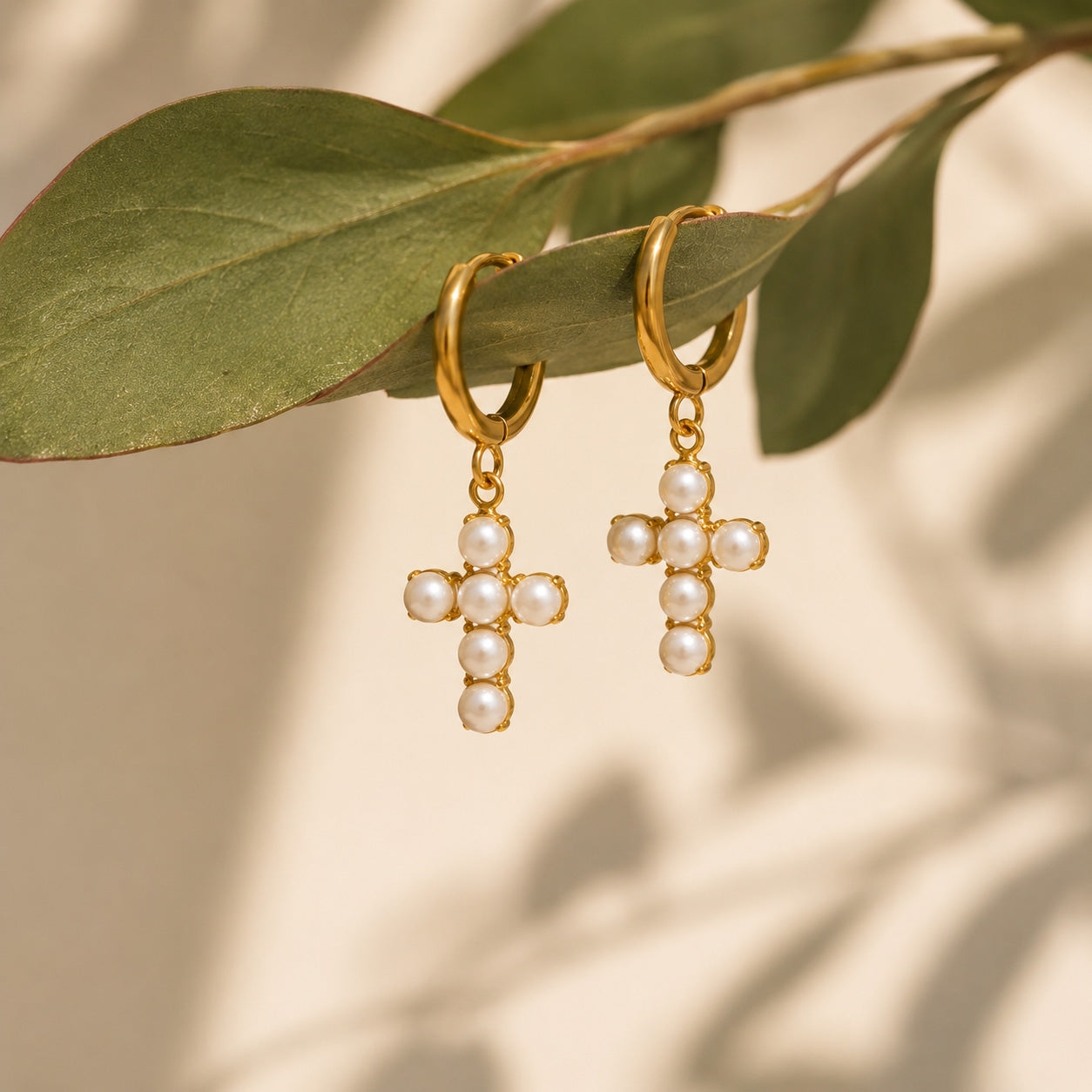 Pair of gold pearl cross drop hoop earrings on cream still life
