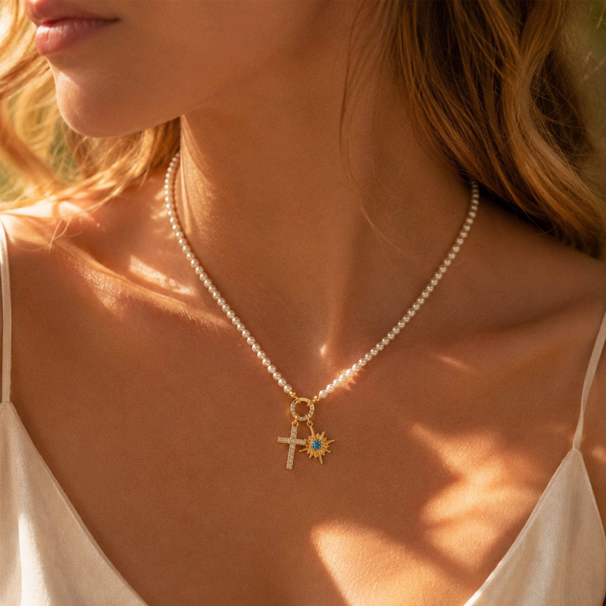 Woman wearing gold pearl necklace with CZ cross charm and blue birthstone sunburst star pendant