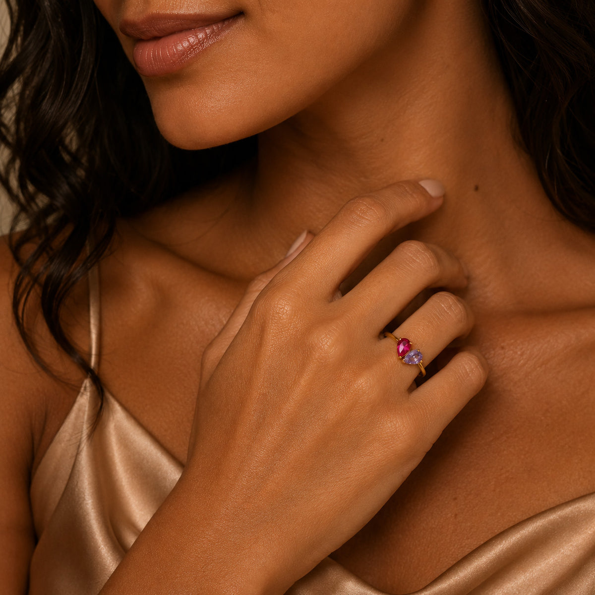 Woman wearing gold toi et moi teardrop birthstone ring with ruby and amethyst stones, hand near collarbone